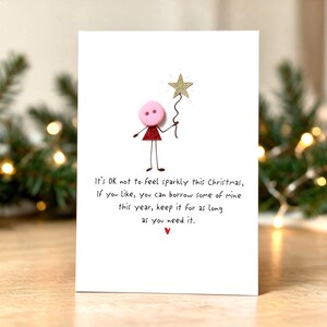Op de afbeelding: Een witte wenskaart met een handgetekend stokfiguurtje met een roze knoop als hoofd en een rood glitterend driehoekig lichaam. Het figuurtje houdt een gouden glitterster vast. De tekst luidt: It's OK not to feel sparkly this Christmas.