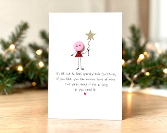 Op de afbeelding: Een witte wenskaart met een handgetekend stokfiguurtje met een roze knoop als hoofd en een rood glitterend driehoekig lichaam. Het figuurtje houdt een gouden glitterster vast. De tekst luidt: It's OK not to feel sparkly this Christmas.