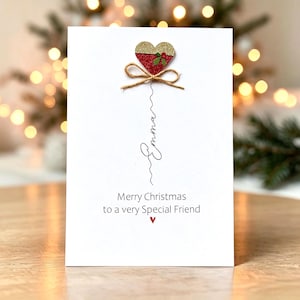 Op de afbeelding: Een witte wenskaart met een glinsterend goud en rood hart met een kleine groene hulsttak en een bruin touwtje als strik. Onder het hart staat de naam Emma in zwarte schrijfletters, gevolgd door de tekst Merry Christmas to a very Special Friend.