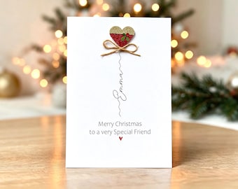 Op de afbeelding: Een witte wenskaart met een glinsterend goud en rood hart met een kleine groene hulsttak en een bruin touwtje als strik. Onder het hart staat de naam Emma in zwarte schrijfletters, gevolgd door de tekst Merry Christmas to a very Special Friend.