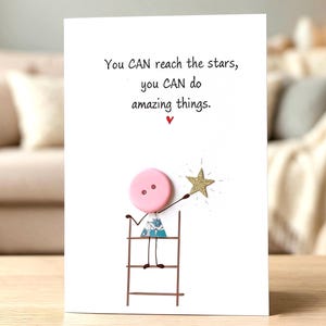 May include: A white greeting card featuring the text You CAN reach the stars, you CAN do amazing things. Below the text is a graphic of a pink button character standing on a brown ladder, reaching toward a gold glittery star with a small red heart.