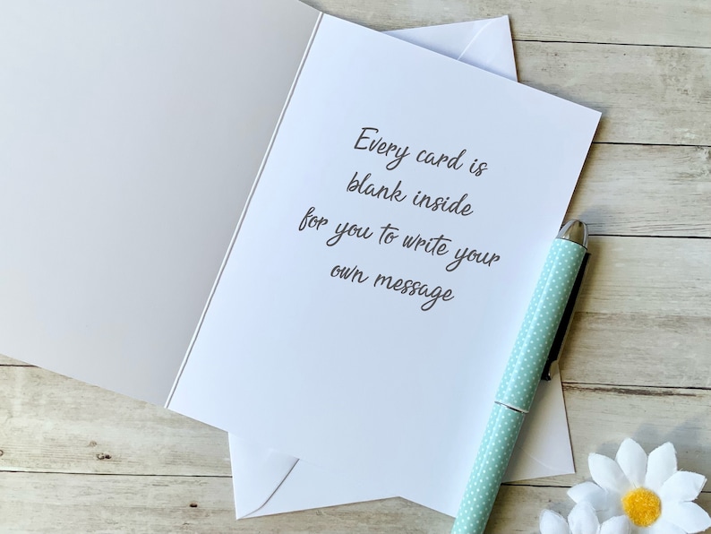 May include: A blank greeting card with the text "Every card is blank inside for you to write your own message". The card is on a wooden surface with a light blue pen and a white daisy.