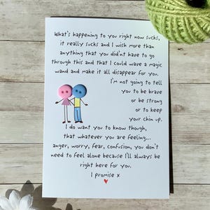 May include: A white greeting card with handwritten text expressing empathy and support. The card features a whimsical illustration of two button figures holding hands. A ball of green yarn sits in the background.
