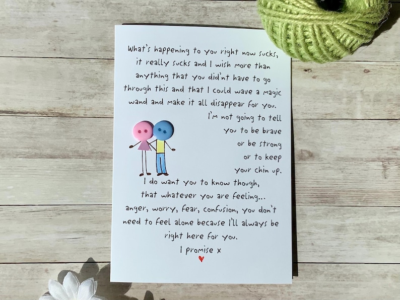 May include: A white greeting card with handwritten text expressing empathy and support. The card features a whimsical illustration of two button figures holding hands. A ball of green yarn sits in the background.