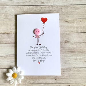 May include: A birthday card with a stick figure holding a red heart balloon. The card features the text "On Your Birthday" and a message of love and support. A white daisy with a yellow center is at the bottom.