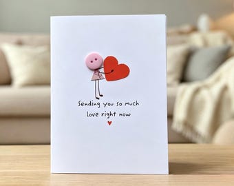 Op de afbeelding: Een witte wenskaart met een illustratie van een stokfiguurtje met een roze knoop als hoofd dat een groot rood hart vasthoudt. Onder het figuurtje staat de tekst "Sending you so much love right now", gevolgd door een klein rood hartsymbool.