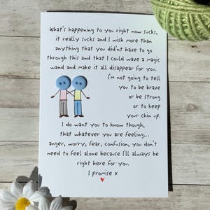 May include: A white greeting card with handwritten text and a button illustration of two figures holding hands. The card's message offers support and comfort. A green ball of twine and a white daisy are in the background.