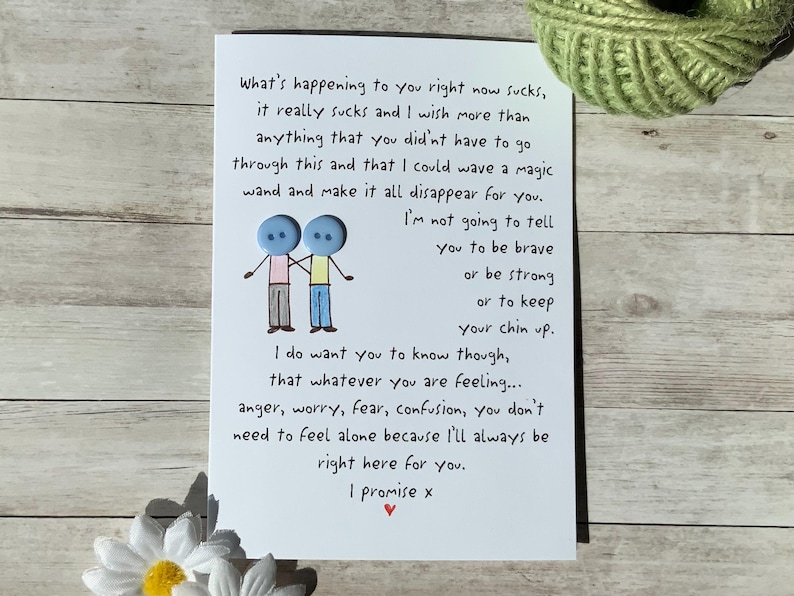 May include: A white greeting card with handwritten text and a button illustration of two figures holding hands. The card's message offers support and comfort. A green ball of twine and a white daisy are in the background.