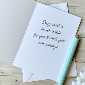 Op de afbeelding: Een blanco kaart met de tekst "Every card is blank inside for you to write your own message". De kaart ligt op een houten oppervlak met een lichtblauwe pen en een witte madeliefje.