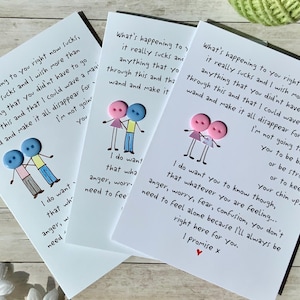 May include: Three white greeting cards with handwritten text and button illustrations. The cards feature a message of support and comfort, with a drawing of two button figures. The cards are on a light wood surface.