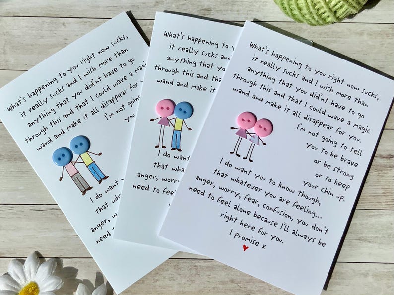 May include: Three white greeting cards with handwritten text and button illustrations. The cards feature a message of support and comfort, with a drawing of two button figures. The cards are on a light wood surface.