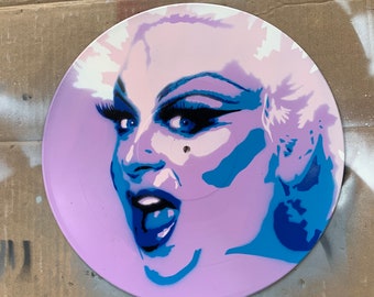 May include: A purple vinyl record with a blue and white stencil of a person's face. The person is wearing makeup and has a surprised expression.