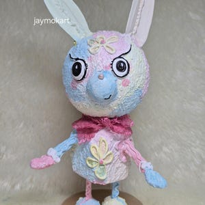 Puede incluir: Una caprichosa figura de conejo en colores pastel, con ojos grandes y orejas largas. El conejo tiene un lazo rosa, una flor en el pecho y está sobre una base redonda. El texto "jaymokart" es visible en la imagen.