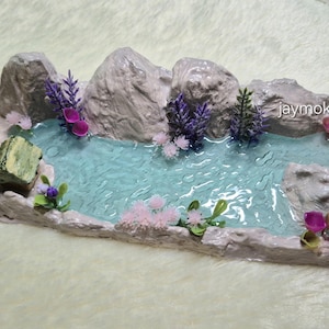 Könnte beinhalten: Miniatur-Zen-Garten-Diorama mit einem ruhigen Teich mit hellblauem Wasser, umgeben von grauen Felsformationen und bunten Miniaturpflanzen. Enthält eine kleine Holzstruktur und einen grünen Stein.