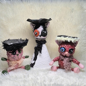 May include: Three handcrafted figurines featuring two small pink cat-like creatures with black hats and a central taller figure with a black and white cone-shaped body, a black hat, and a face painted with a large orange eye and a black eye patch.