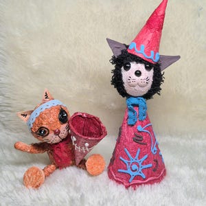 May include: Two handcrafted cat figurines. One is a seated orange cat with a light blue headband holding a dark red cone. The other is a standing cat wearing a tall pink pointed hat with blue swirls, a blue scarf, and a pink cone-shaped body.
