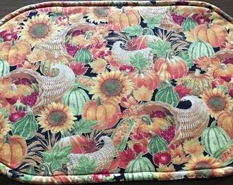 May include: Oval-shaped placemat with a fall harvest theme. The fabric features pumpkins, sunflowers, gourds, cornucopias, and autumn leaves in shades of orange, yellow, green, and red against a dark background. The edges are finished with a stitched border.
