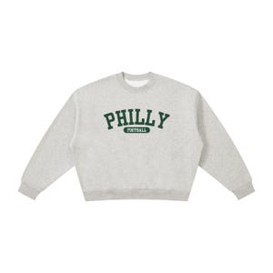 Könnte beinhalten: Ein hellgrau meliertes Sweatshirt mit Rundhalsausschnitt, das auf der Brust das Wort PHILLY in einer dunkelgrünen, bogenförmigen Schrift im College-Stil zeigt, mit dem Wort FOOTBALL in einem kleinen dunkelgrünen Kasten direkt darunter.