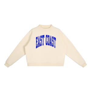 Puede incluir: Una sudadera de manga larga de color crema con cuello alto y puños acanalados. La parte delantera de la prenda muestra el texto "EAST COAST" en una fuente azul, arqueada y de estilo universitario con una textura desgastada.