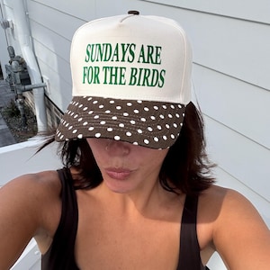 May include: A cream-colored baseball cap featuring the green text "SUNDAYS ARE FOR THE BIRDS" on the front panel. The cap has a dark brown brim decorated with a white polka dot pattern and a small dark brown button at the top center of the crown.