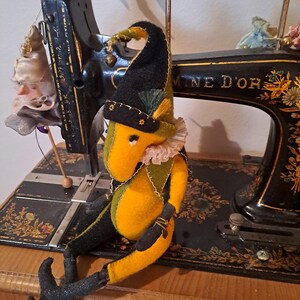 Könnte beinhalten: Handgefertigte Filz-Narrenpuppe mit gelbem Körper, schwarzen und grünen Akzenten und einem schwarzen Hut mit goldenem Besatz. Die Puppe sitzt auf einer alten schwarzen Nähmaschine mit goldenen Blumenmustern. Eine kleine Puppe hängt an der Maschine.