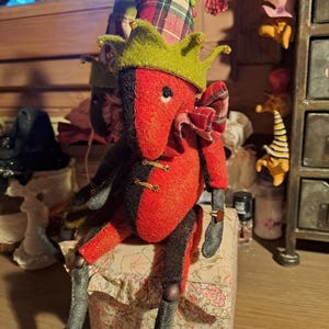 Könnte beinhalten: Ein rotes und schwarzes Filzspielzeug mit einem grün-rot karierten Hut, das auf einer Schachtel mit Blumenmuster sitzt. Das Spielzeug hat eine lange Nase und eine Schleife.