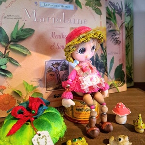 Könnte beinhalten: Eine kleine Puppe mit einem rosa und gelben Häkelhut sitzt auf einer alten Blechdose. Die Puppe hat braune Augen und ein rosa Kleid. Die Puppe ist umgeben von grünen Pflanzen, einem grün-roten Pilz und einem kleinen Frosch.