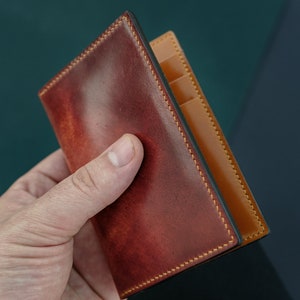 Peut inclure: Un portefeuille en cuir marron avec un int&eacute;rieur en cuir beige et des coutures jaunes. Le portefeuille est pli&eacute; en deux et poss&egrave;de plusieurs fentes pour cartes.