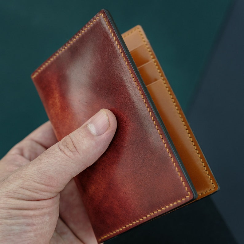 Peut inclure: Un portefeuille en cuir marron avec un int&eacute;rieur en cuir beige et des coutures jaunes. Le portefeuille est pli&eacute; en deux et poss&egrave;de plusieurs fentes pour cartes.