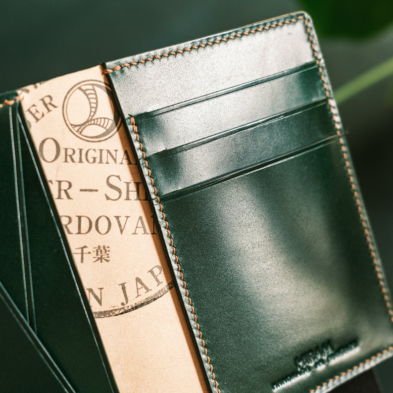 Peut inclure: Un portefeuille en cuir vert avec un bord cousu et un rabat en cuir marron. Le rabat porte le texte "Original Leather - Shell Cordovan" et "干葉" en japonais. Le portefeuille a plusieurs emplacements pour cartes.