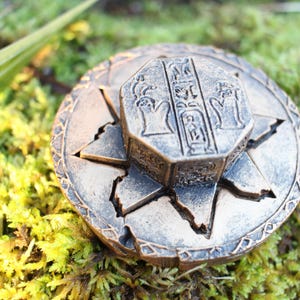 May include: A bronze-colored, circular decorative object with an octagonal centerpiece. The centerpiece features hieroglyphic-style carvings. The object is set against a backdrop of green moss and foliage, suggesting an ancient or historical theme.