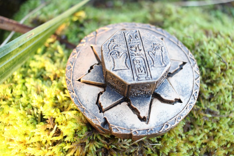 May include: A bronze-colored, circular decorative object with an octagonal centerpiece. The centerpiece features hieroglyphic-style carvings. The object is set against a backdrop of green moss and foliage, suggesting an ancient or historical theme.