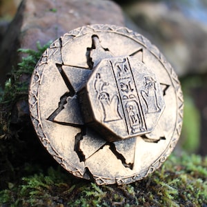 May include: A bronze-colored, circular pendant with an Egyptian hieroglyphic design. The pendant features a star-shaped outer ring and a central, octagonal piece with detailed carvings. The pendant is resting on a bed of moss and stone.