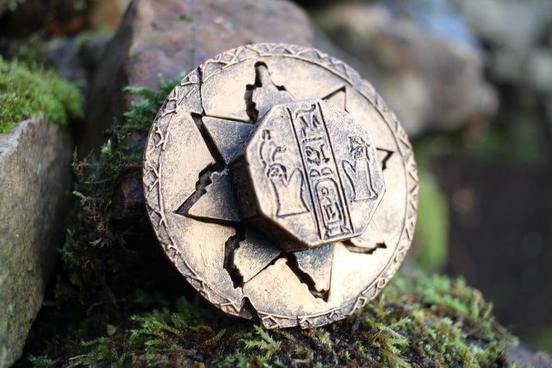 May include: A bronze-colored, circular pendant with an Egyptian hieroglyphic design. The pendant features a star-shaped outer ring and a central, octagonal piece with detailed carvings. The pendant is resting on a bed of moss and stone.