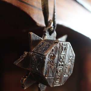 Puede incluir: Un colgante de metal plateado con detalles ornamentados, con forma de estrella y un centro hexagonal. El colgante est&aacute; colgado de una cinta negra.