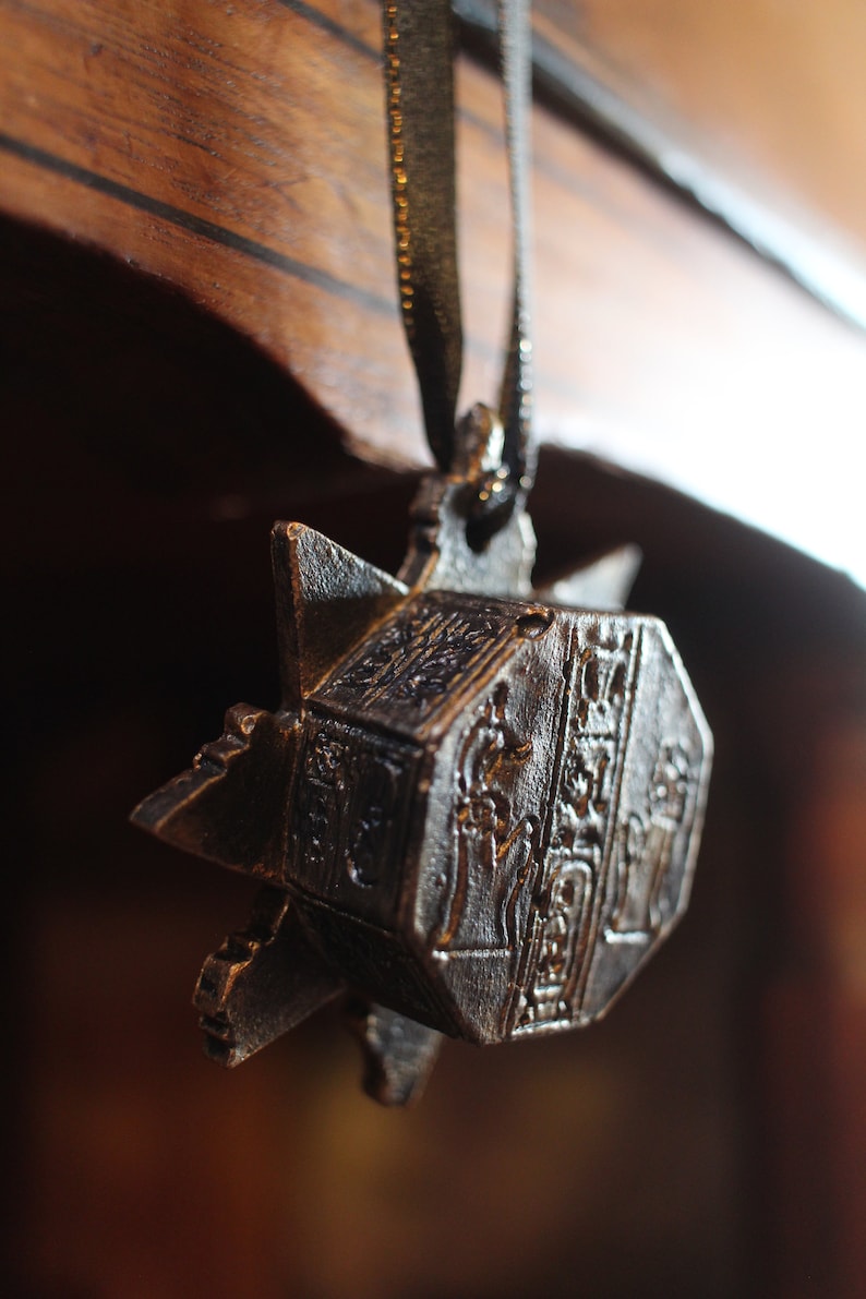 Puede incluir: Un colgante de metal plateado con detalles ornamentados, con forma de estrella y un centro hexagonal. El colgante est&aacute; colgado de una cinta negra.