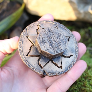 May include: A bronze-colored, round puzzle box with an octagonal center piece. The box features star-shaped cutouts and hieroglyphic-style engravings. The box is held in a hand against a backdrop of greenery and moss.