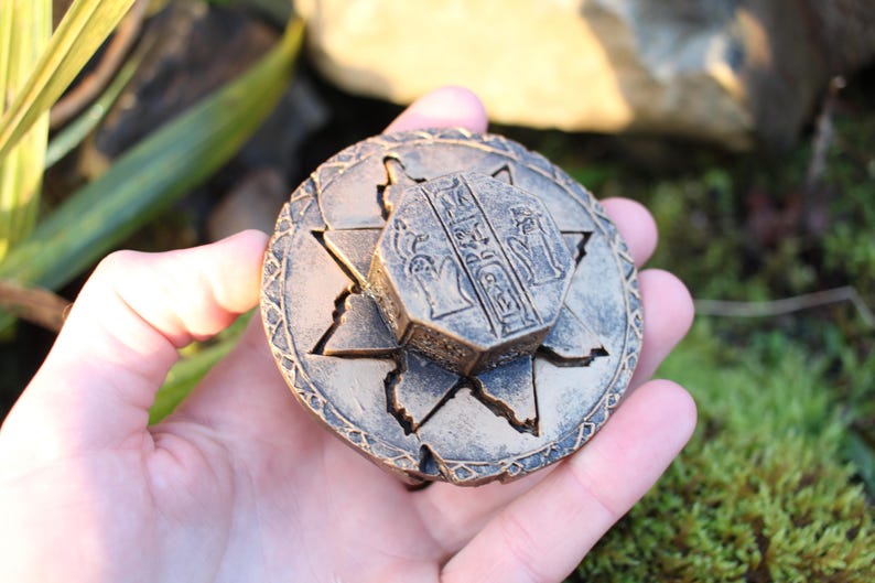 May include: A bronze-colored, round puzzle box with an octagonal center piece. The box features star-shaped cutouts and hieroglyphic-style engravings. The box is held in a hand against a backdrop of greenery and moss.