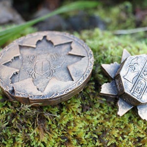 May include: Two bronze-colored, decorative objects rest on a bed of green moss. One is a round medallion with a starburst design and a scarab beetle. The other is a star-shaped object with hieroglyphic carvings.