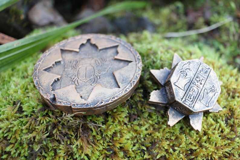 May include: Two bronze-colored, decorative objects rest on a bed of green moss. One is a round medallion with a starburst design and a scarab beetle. The other is a star-shaped object with hieroglyphic carvings.
