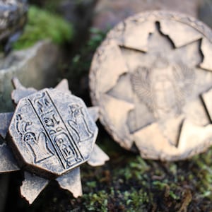 May include: Two bronze-colored, ancient-style medallions. One is a starburst shape with hieroglyphs, the other is round with a starburst design and an emblem. The medallions are on a mossy surface.