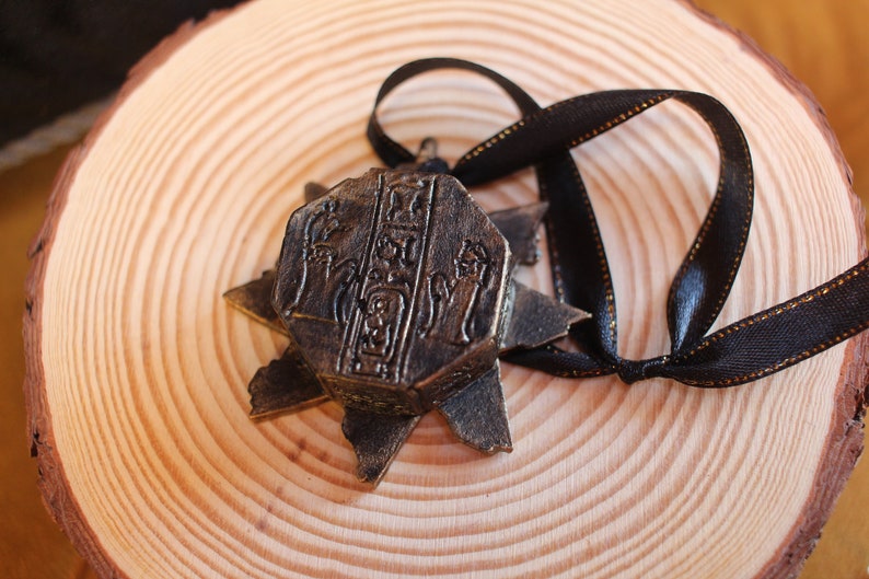 Puede incluir: Un colgante de estrella de color bronce con tallas intrincadas y una cinta negra. El colgante est&aacute; sobre una superficie de madera.