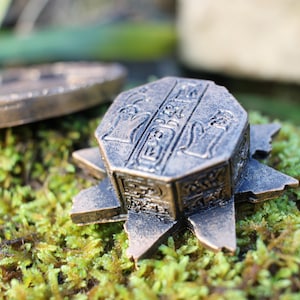 May include: Bronze-colored, star-shaped decorative box with hieroglyphic carvings. The box sits on a bed of green moss, with a circular object in the background. The box has an aged, antique appearance.