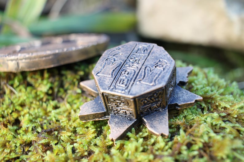 May include: Bronze-colored, star-shaped decorative box with hieroglyphic carvings. The box sits on a bed of green moss, with a circular object in the background. The box has an aged, antique appearance.