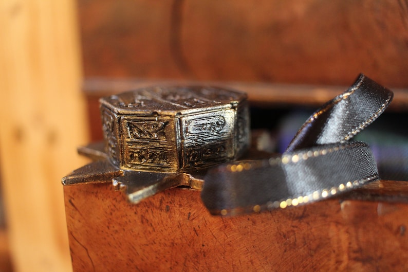 Puede incluir: Un adorno de estrella de metal dorado con una pieza central octogonal detallada y de m&uacute;ltiples capas. El adorno est&aacute; sobre una superficie de madera con una cinta negra.
