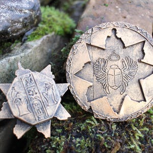 May include: Two bronze-colored, ancient-style medallions. One is a starburst shape with hieroglyphs, the other is round with a scarab beetle and wings design. The medallions are set against a backdrop of rocks and moss.