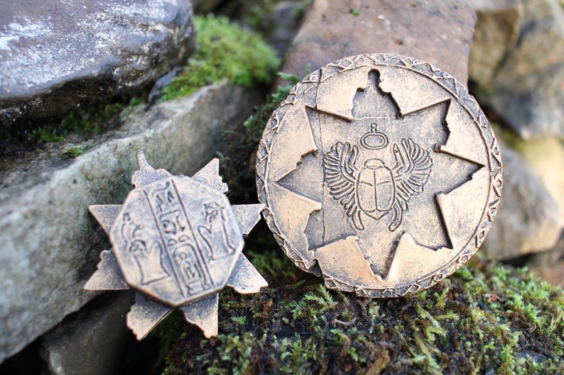 May include: Two bronze-colored, ancient-style medallions. One is a starburst shape with hieroglyphs, the other is round with a scarab beetle and wings design. The medallions are set against a backdrop of rocks and moss.