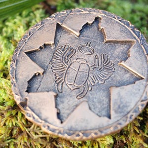 May include: A bronze-colored round coin with an embossed scarab beetle design inside a star-shaped cutout. The coin has a detailed border and is set against a backdrop of green moss.