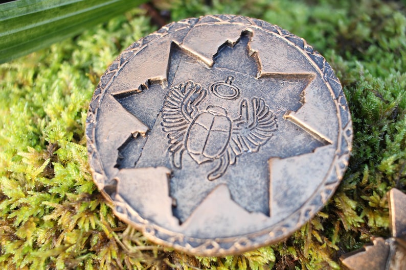 May include: A bronze-colored round coin with an embossed scarab beetle design inside a star-shaped cutout. The coin has a detailed border and is set against a backdrop of green moss.