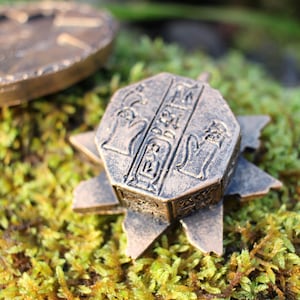 May include: Bronze-colored, octagonal pendant with hieroglyphic carvings and a star-shaped base. Another bronze coin is visible in the background, all resting on a bed of green moss. The pendant has a detailed, aged appearance.